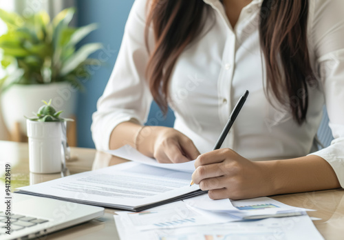 Focused Businesswoman Writing in an Office Setting. A professional woman with long brown hair and light skin working diligently at a desk, surrounded by documents and a laptop. 