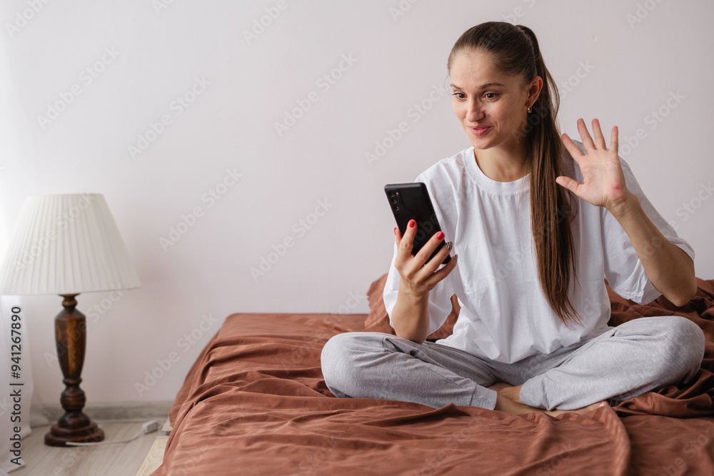 Happy smiling woman is making a video call using a cellphone while sitting on the bed Happy smiling woman is making a video call using a cellphone while sitting on the bed