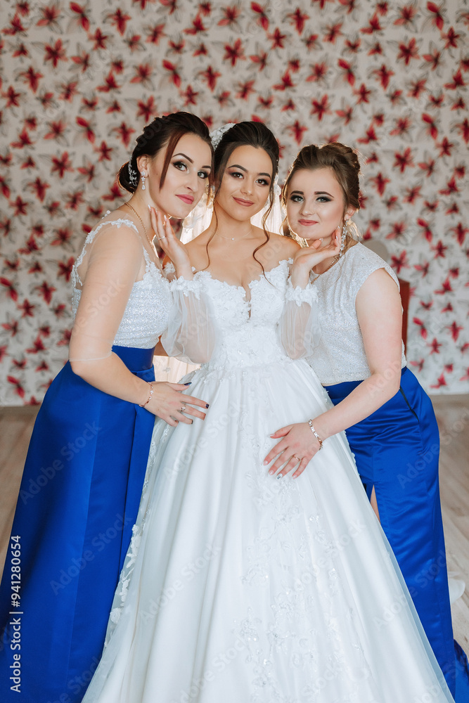 Three women are posing for a picture in front of a wall with red flowers. The bride is wearing a white dress and the other two women are wearing blue dresses. Scene is happy and celebratory Three women are posing for a picture in front of a wall with red flowers. The bride is wearing a white dress and the other two women are wearing blue dresses. Scene is happy and celebratory