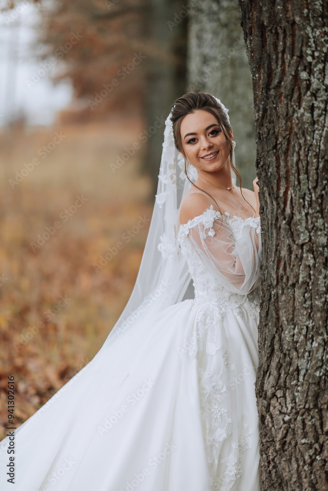 A woman in a white wedding dress is standing behind a tree. She is smiling and looking at the camera A woman in a white wedding dress is standing behind a tree. She is smiling and looking at the camera