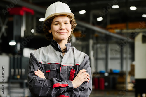 Portrait of confident female construction worker wearing hard hat and industrial uniform standing inside factory photo