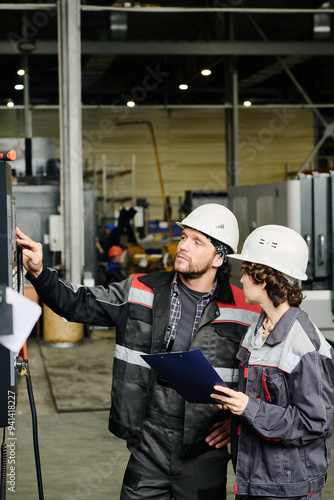 Workers wearing protective gear and hard hats, examining machinery inside factory, discussing maintenance procedures photo