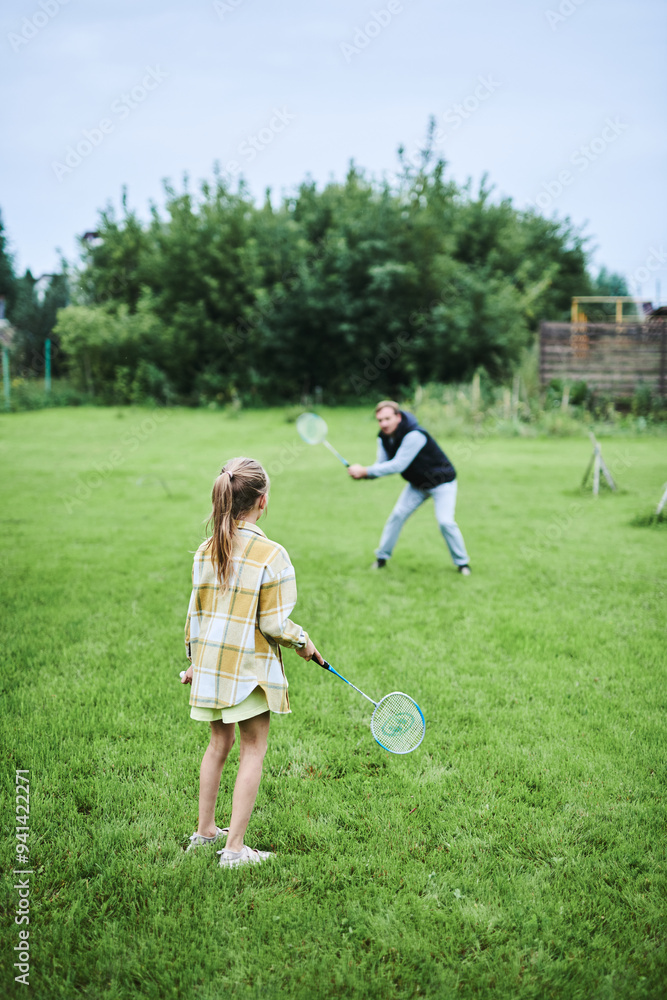 Fototapeta premium Teenage girl playing badminton with her father on green grass in summer park