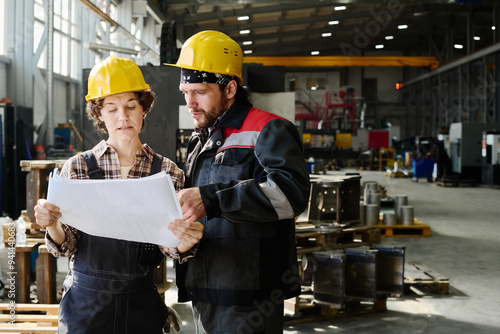 Two factory workers in hard hats examining paper blueprints indoors in manufacturing facility surrounded by machinery and metal parts