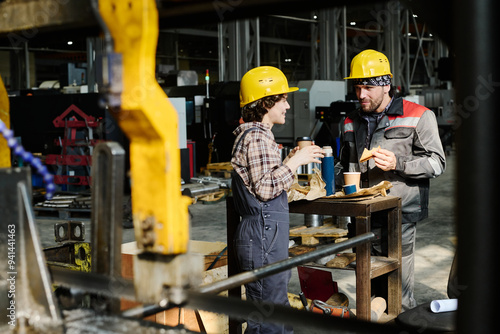 Two industrial workers wearing safety helmets and holding coffee cups, discussing during a break photo