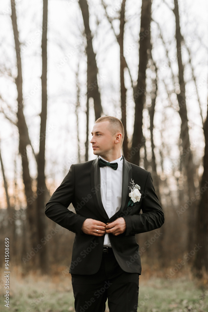 A man in a black suit and bow tie stands in a field of trees. He is adjusting his tie and holding a flower in his hand. Concept of elegance and sophistication, as the man is dressed in formal attire A man in a black suit and bow tie stands in a field of trees. He is adjusting his tie and holding a flower in his hand. Concept of elegance and sophistication, as the man is dressed in formal attire