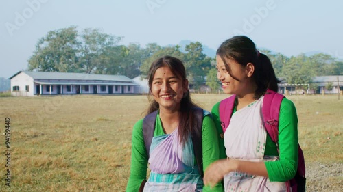 Two young school bound Assamese Indian females or girls stand in the rural landscape outside a school in uniforms smile looking at or facing the camera. Education, learning, gender equality concept
