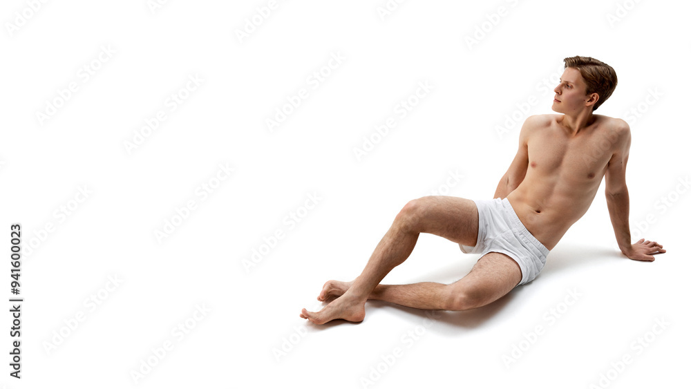 Fototapeta premium Shirtless man in white boxers relaxes on floor with one leg extended, leaning back on his hands, looking off into distance with calm expression. Concept of male health, natural beauty, fashion, care.