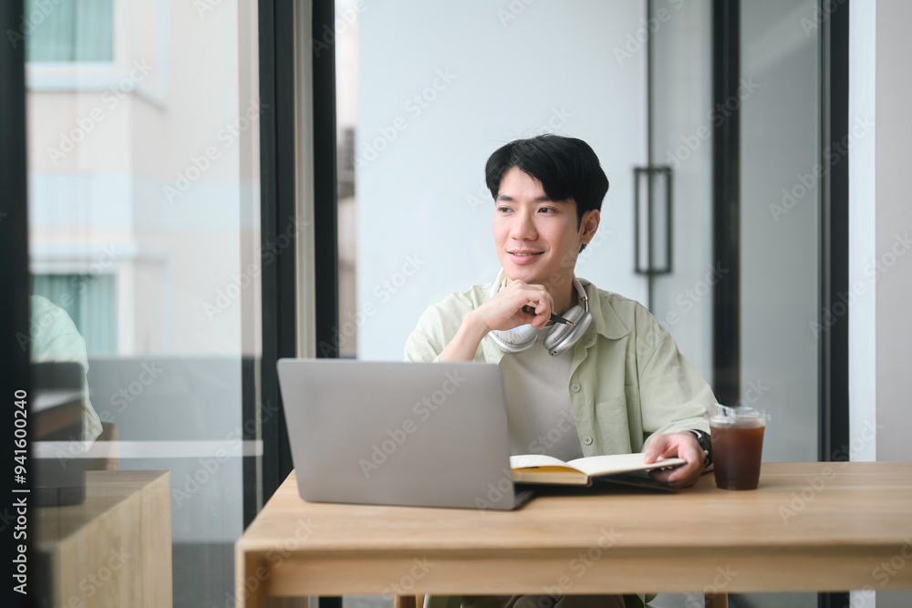 A young man sits at a desk with a laptop and a book. He is wearing headphones and he is deep in thought A young man sits at a desk with a laptop and a book. He is wearing headphones and he is deep in thought