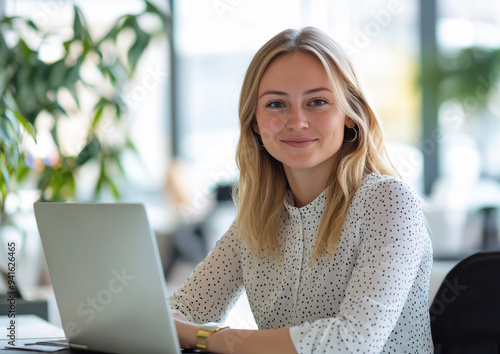 A confident businesswoman smiles while productively working on her laptop in a bright, modern office space.