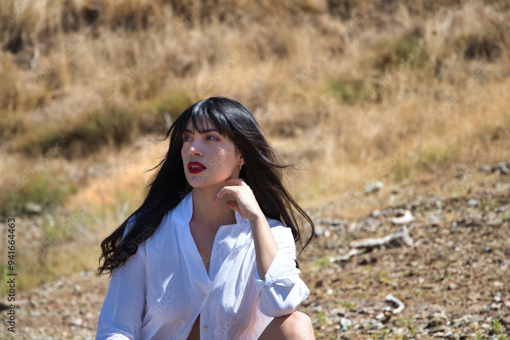 Young, dark, young and beautiful latin woman dressed in white shirt and black shorts and straw hat sitting on a rock in a field. The woman rests her chin on her hand to make herself comfortable. Young, dark, young and beautiful latin woman dressed in white shirt and black shorts and straw hat sitting on a rock in a field. The woman rests her chin on her hand to make herself comfortable.