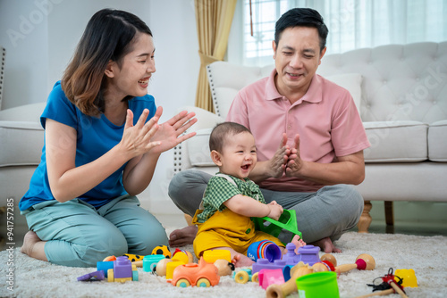 A family of three, a man and two women, are playing with toys on the floor. The baby is holding a toy truck and the parents are clapping. Scene is happy and playful
