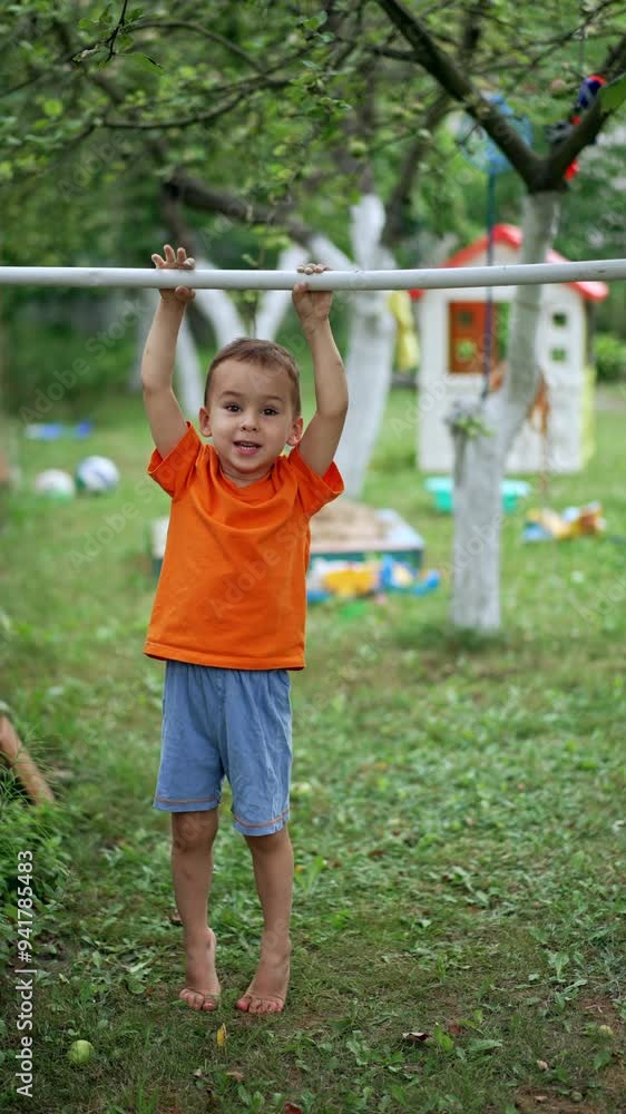 Beautiful Caucasian kid training on the horizontal bar. Toddler doing sport since early age. Garden at backdrop. Vertical video. Beautiful Caucasian kid training on the horizontal bar. Toddler doing sport since early age. Garden at backdrop. Vertical video.