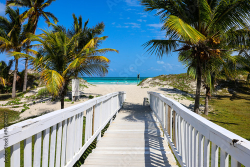 Fototapeta Naklejka Na Ścianę i Meble -  Picturesque view to tropical beach with white sand and coconut palm trees. Wooden path to ocean coast, tourist resort on Caribbean island