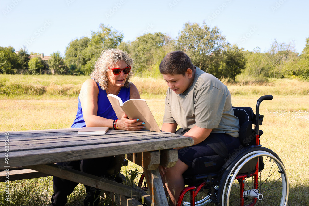 Boy in wheelchair reading a book with woman outdoors Boy in wheelchair reading a book with woman outdoors