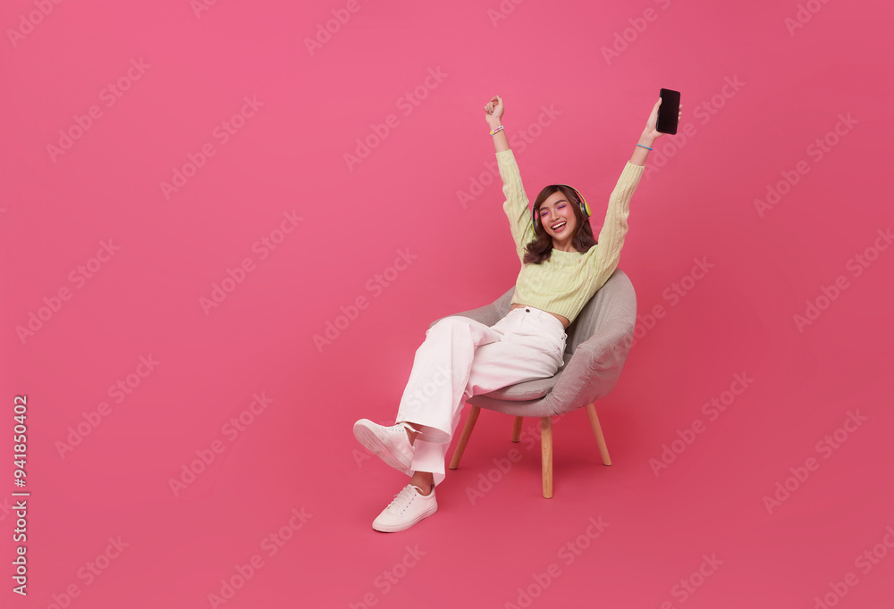 Cheerful young asian teen woman in wireless headphones sitting in armchair, listening to music, raising her arm, dancing to favorite song, enjoying popular soundtrack isolated on pink background.