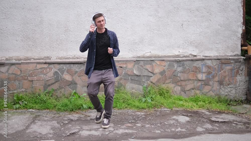 young man in blue checkered shirt and black t-shirt listens to music with large white headphones and dance. He stands outdoors against light-colored textured wall with green leafy plants background young man in blue checkered shirt and black t-shirt listens to music with large white headphones and dance. He stands outdoors against light-colored textured wall with green leafy plants background