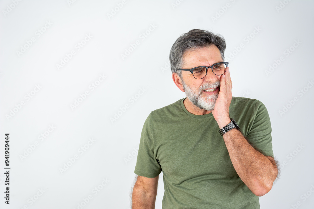 Obraz premium A concerned elderly man in a casual green t-shirt standing isolated, touching his cheek due to a toothache, with a pained expression. Senior hispanic man with beard wearing casual clothes over grey