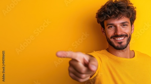 A confident man dressed in a yellow shirt smiles warmly while pointing towards the viewer, exuding a sense of assurance and positivity against a matching vibrant yellow background. photo