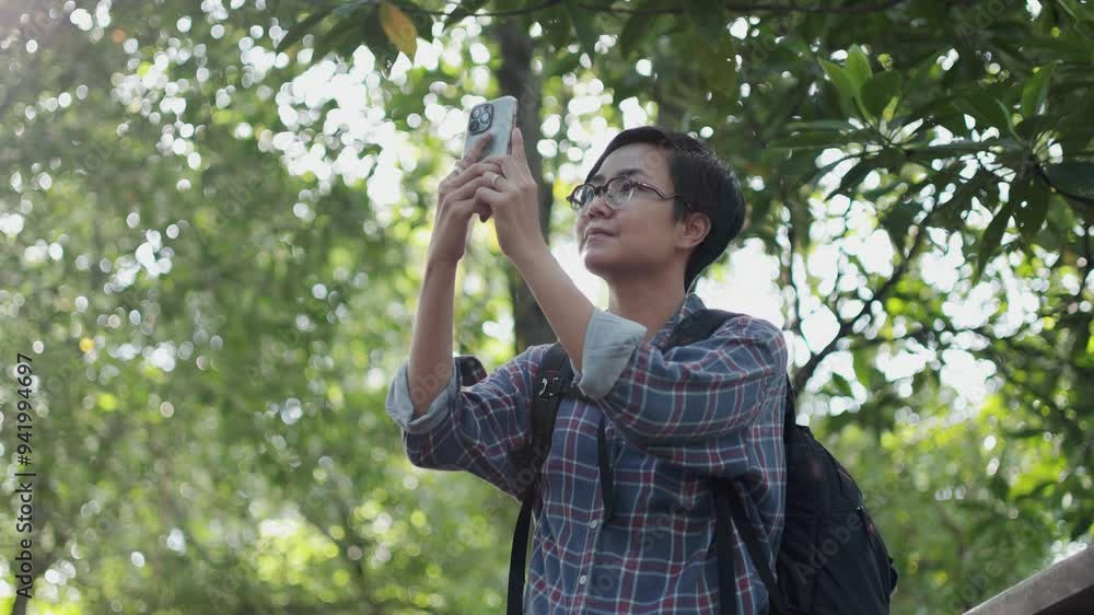 A young, confident Asian woman with short hair is walking through a lush forest with taking photo from her smartphone. She is enjoying the serenity and greenery with freedom of the nature outdoors. A young, confident Asian woman with short hair is walking through a lush forest with taking photo from her smartphone. She is enjoying the serenity and greenery with freedom of the nature outdoors.