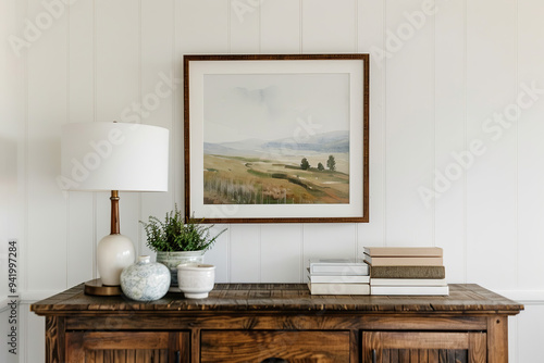 A large framed landscape painting hanging on the wall of a farmhouse, above a rustic wood sideboard with books and white lamps, in neutral tones with pops of greenery, farmhouse-style home decor photo
