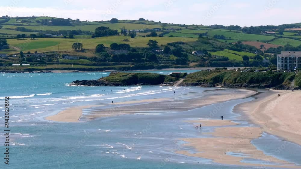 A wide view of a shoreline with a beach and a small river running inland. The water is turquoise blue and calm, and there are a few people walking on the sand. A wide view of a shoreline with a beach and a small river running inland. The water is turquoise blue and calm, and there are a few people walking on the sand.