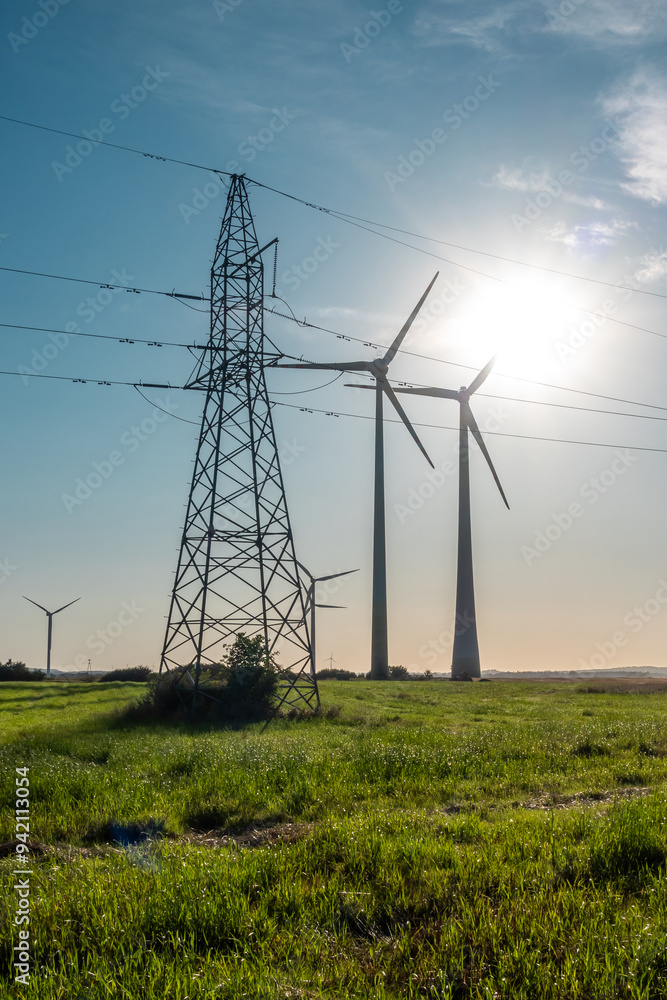 Wind turbines and high voltage electric pylon against blue sky. Sun shining behind the turbines