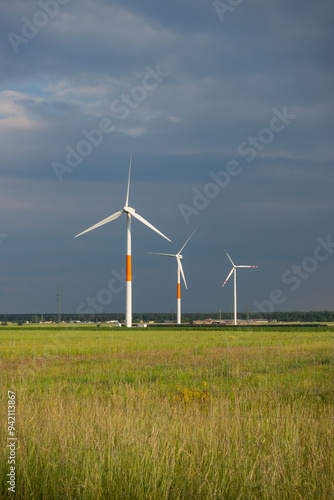 Obraz na płótnie Distant white wind turbines against a dramatic cloudy sky. Meadow in the foreground, taken late afternoon w salonie