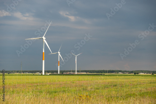 Obraz na płótnie Distant white wind turbines against a dramatic cloudy sky. Meadow in the foreground, taken late afternoon w salonie