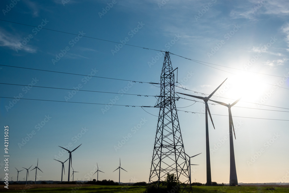 Wind turbines and high voltage electric pylon against blue sky. Sun shining behind the turbines