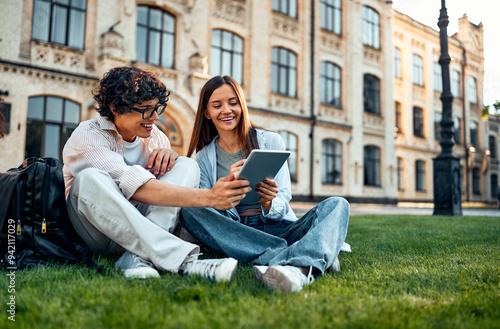 Two young students are sitting on the grass on the campus talking and preparing for classes. Real student life.
