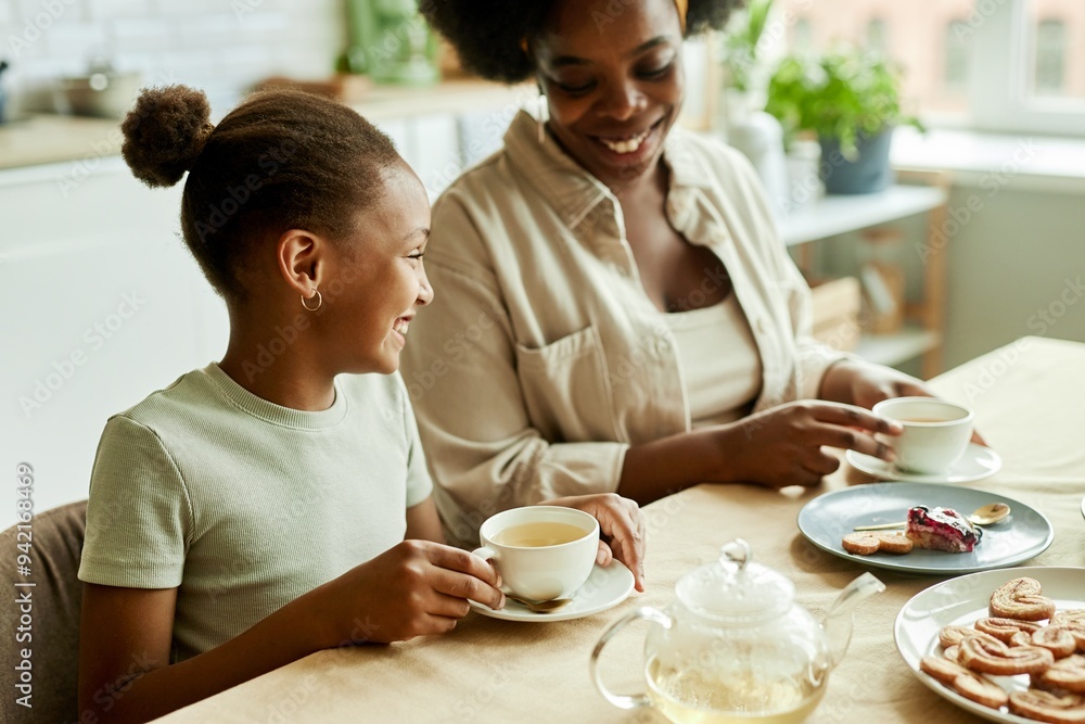Side view of smiling African American little daughter cheerfully talking to young mother, while having tea with sweet treat together at family gathering at kitchen table Side view of smiling African American little daughter cheerfully talking to young mother, while having tea with sweet treat together at family gathering at kitchen table
