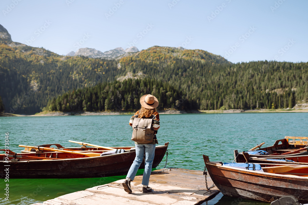 A woman with curly hair poses by the lake on a sunny day, wearing a colorful sweater and a wide-brimmed hat while nearby wooden boats are docked. Mountains, Lake, green forests A woman with curly hair poses by the lake on a sunny day, wearing a colorful sweater and a wide-brimmed hat while nearby wooden boats are docked. Mountains, Lake, green forests