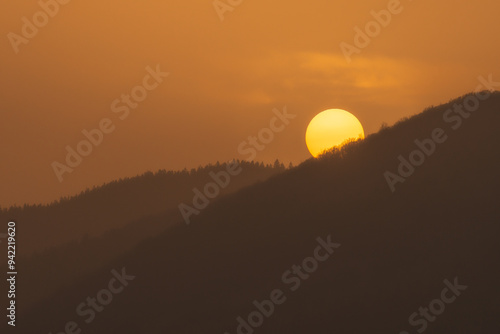 Sunset over the mountains in the Sudetes, the orange sun hides behind the dense forest on the hill. photo