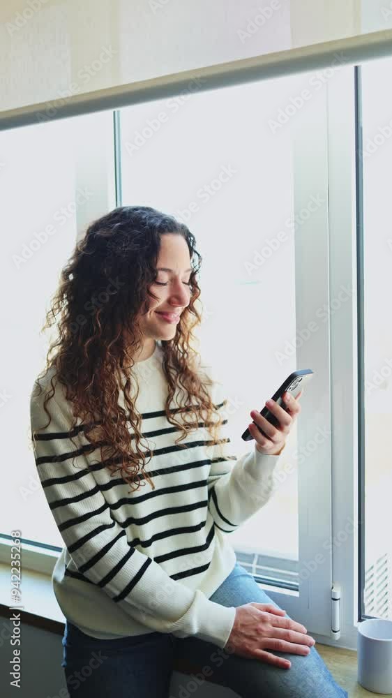 Woman standing by the window, holding a smartphone and smiling, enjoys a moment of connection and relaxation indoors. Woman smiling and using smartphone by the window Woman standing by the window, holding a smartphone and smiling, enjoys a moment of connection and relaxation indoors. Woman smiling and using smartphone by the window