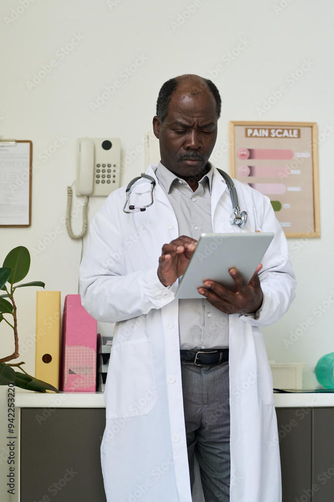 Middle-aged doctor wearing white coat using a tablet while standing in medical office with pain scale chart on wall and various office items around Middle-aged doctor wearing white coat using a tablet while standing in medical office with pain scale chart on wall and various office items around