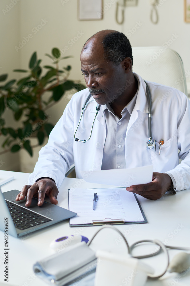 Senior doctor working in medical office, viewing patient records on laptop, reading medical chart with stethoscope draped around neck, showing serious expression Senior doctor working in medical office, viewing patient records on laptop, reading medical chart with stethoscope draped around neck, showing serious expression