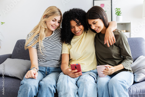 Three young multiracial women watching social media content on smart phone app sitting on sofa at home. Technology and friendship concept photo