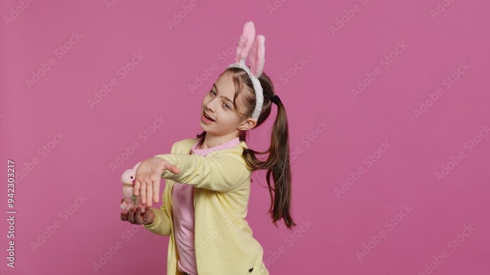Lovely little girl with bunny ears sending air kisses in studio, presenting a stuffed pink rabbit toy for easter holiday. Smiling energetic preteen feeling positive and carefree. Camera A. Lovely little girl with bunny ears sending air kisses in studio, presenting a stuffed pink rabbit toy for easter holiday. Smiling energetic preteen feeling positive and carefree. Camera A.