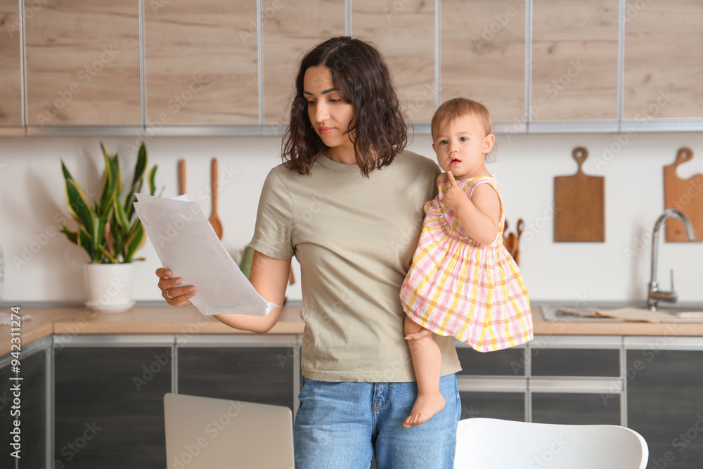 Cute little baby and young mother with laptop reading documents on maternity leave in kitchen Cute little baby and young mother with laptop reading documents on maternity leave in kitchen