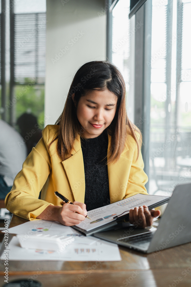 A young woman in a yellow blazer working at a desk in a modern office, writing in a notebook with a laptop and documents in front of her. A young woman in a yellow blazer working at a desk in a modern office, writing in a notebook with a laptop and documents in front of her.