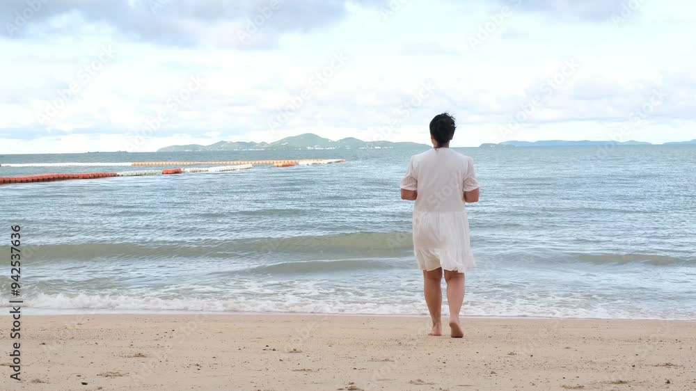 mid aged woman in white summer clothes walking on the seashore on sandy beach, holding smartphone in hands mid aged woman in white summer clothes walking on the seashore on sandy beach, holding smartphone in hands