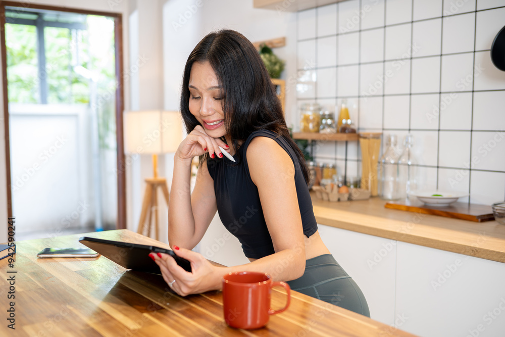 Fototapeta premium An attractive Asian woman is using her digital tablet at the kitchen table in her minimalist kitchen