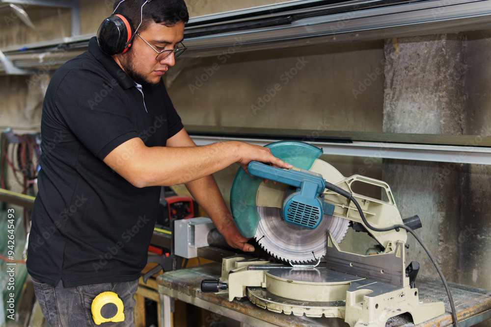 A man is using a power saw to cut a piece of wood. He is wearing a black shirt and a pair of headphones