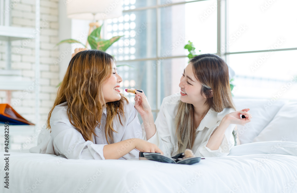 Two happy beautiful Asian female smile lying relaxed on a white bedroom, putting makeup. Attractive woman having fun together trying beauty products, Concept teenage or girlfriends lifestyle at home. Two happy beautiful Asian female smile lying relaxed on a white bedroom, putting makeup. Attractive woman having fun together trying beauty products, Concept teenage or girlfriends lifestyle at home.