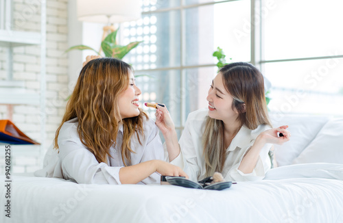 Two happy beautiful Asian female smile lying relaxed on a white bedroom, putting makeup. Attractive woman having fun together trying beauty products, Concept teenage or girlfriends lifestyle at home.