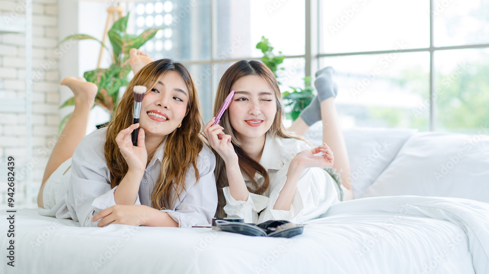 Two happy beautiful Asian female smile lying relaxed on a white bedroom, putting makeup. Attractive woman having fun together trying beauty products, Concept teenage or girlfriends lifestyle at home. Two happy beautiful Asian female smile lying relaxed on a white bedroom, putting makeup. Attractive woman having fun together trying beauty products, Concept teenage or girlfriends lifestyle at home.