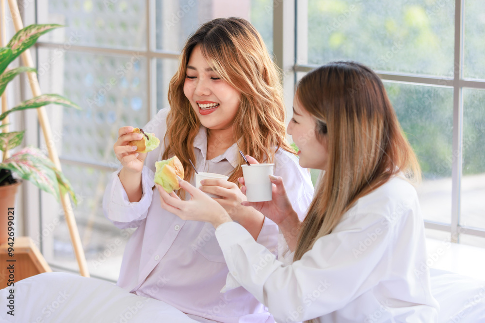 Two beautiful people happy Asian females drinking hot coffee or tea with bread or cake. Pretty woman laughing and talking relaxed funny on a white bed, Concept teenage or friends lifestyle at home. Two beautiful people happy Asian females drinking hot coffee or tea with bread or cake. Pretty woman laughing and talking relaxed funny on a white bed, Concept teenage or friends lifestyle at home.
