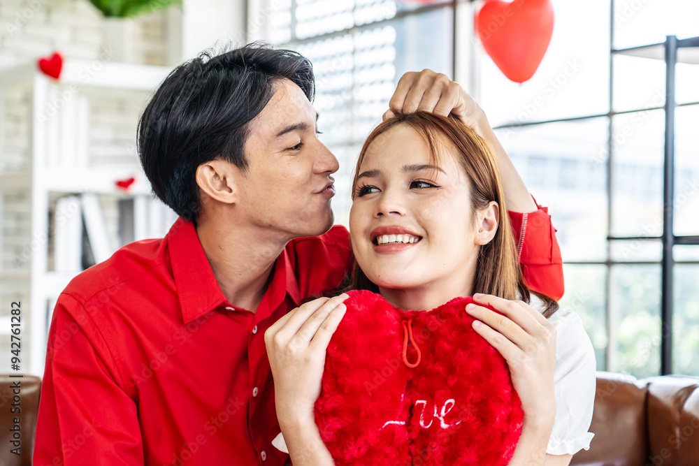 Portrait of asian young couple sitting on sofa smiling and holding red heart pillow in living room for romance and celebrating valentine day. Concept love story of young couple in valentine's day. Portrait of asian young couple sitting on sofa smiling and holding red heart pillow in living room for romance and celebrating valentine day. Concept love story of young couple in valentine's day.