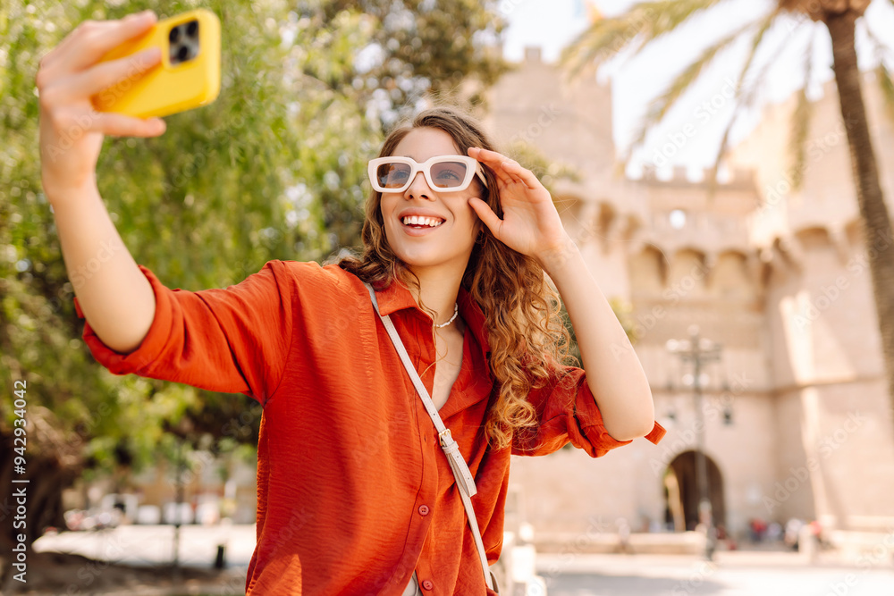 Cheerful woman taking a selfie in stylish sunglasses while strolling down a sunlit city street during daytime. Travel, tourism, technology, blogging, communication concept. Cheerful woman taking a selfie in stylish sunglasses while strolling down a sunlit city street during daytime. Travel, tourism, technology, blogging, communication concept.